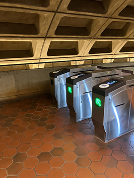 A picture of the entrance of the guided pathway next to the faregates. To the left there is a yellow wet floor cone next to a faregate and to the right in the background there is a sign pointing to the elevator on the wall. The wall in the background has a railing on top of it.